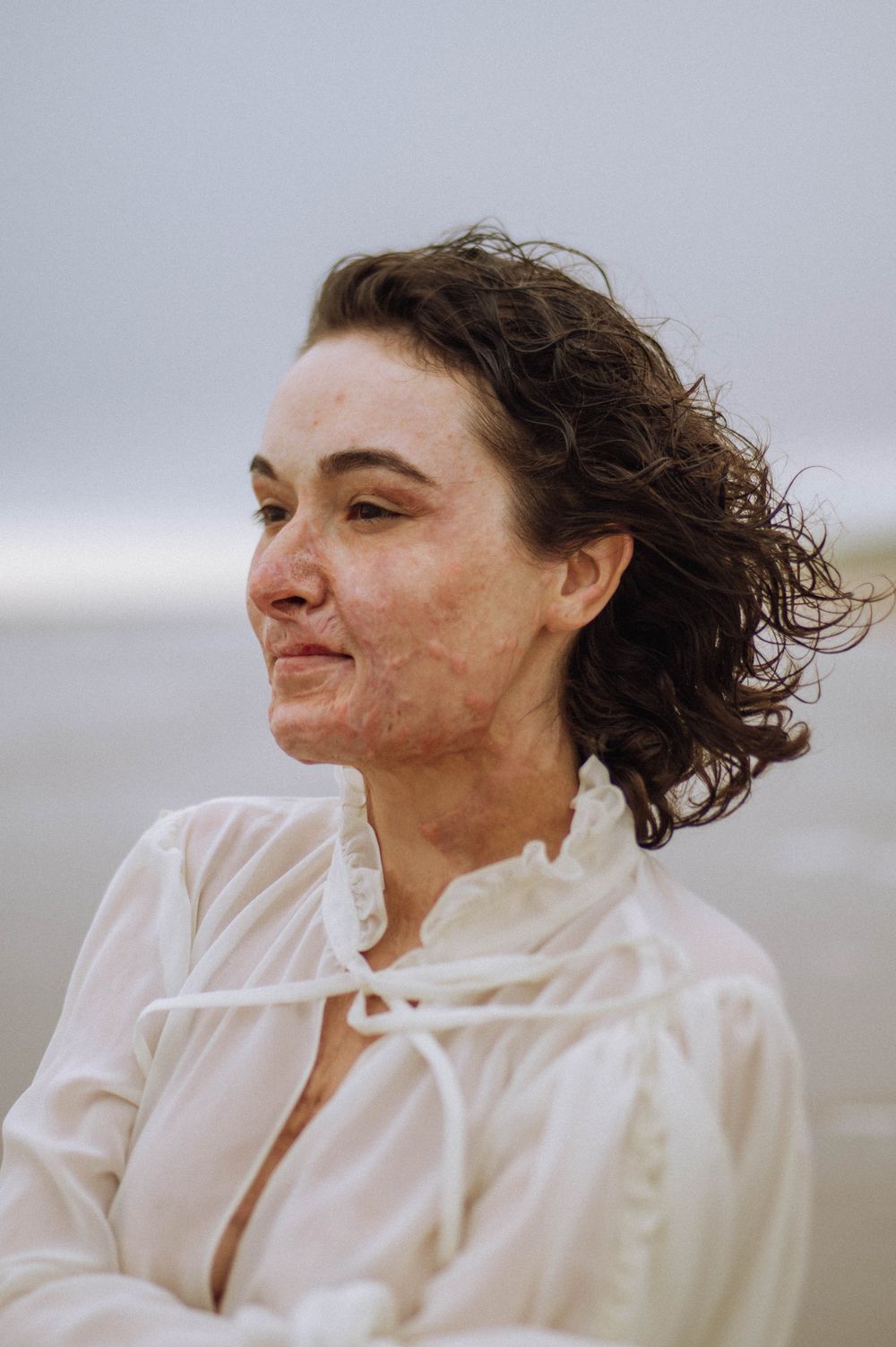 Person in flowing white dress stands on windswept beach.