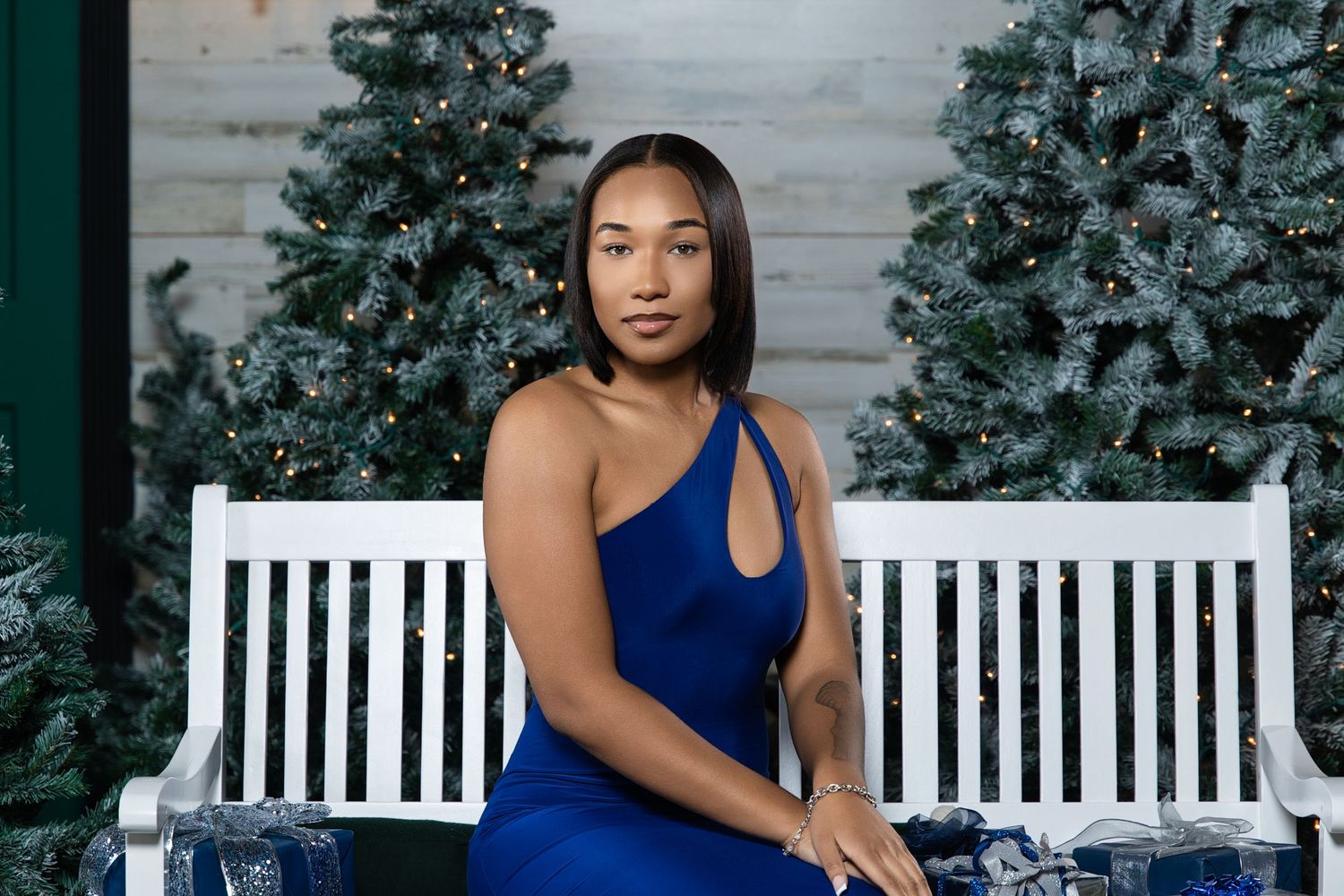 Woman in blue dress poses on white bench with decorated Christmas trees in background.