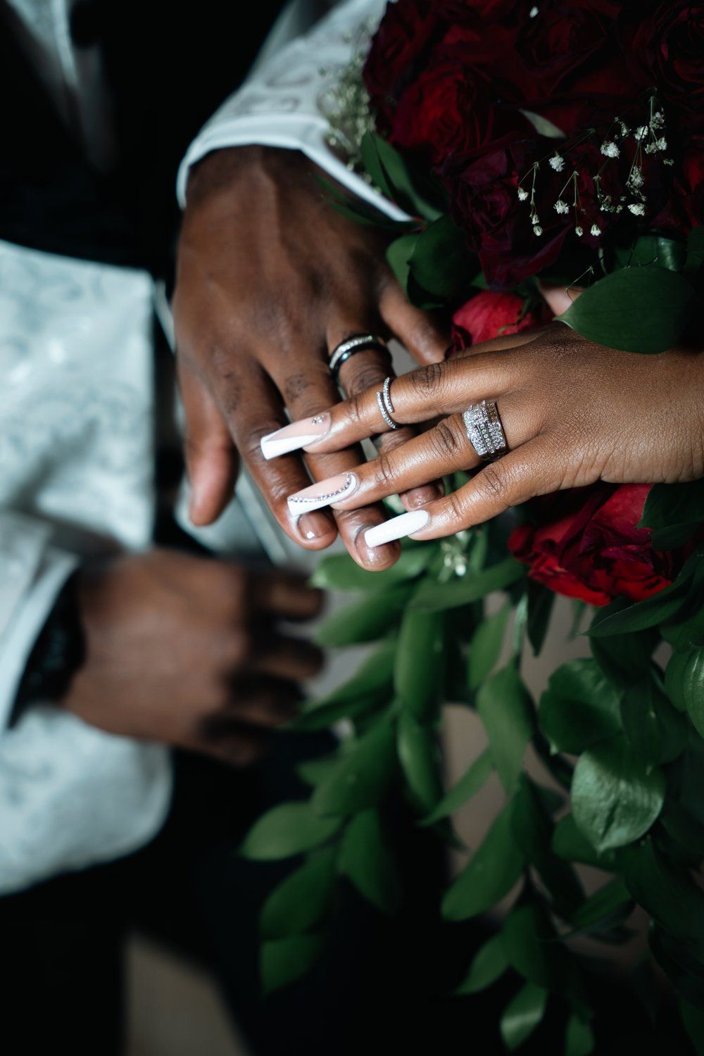 Close-up of hands with wedding rings holding red roses and greenery.