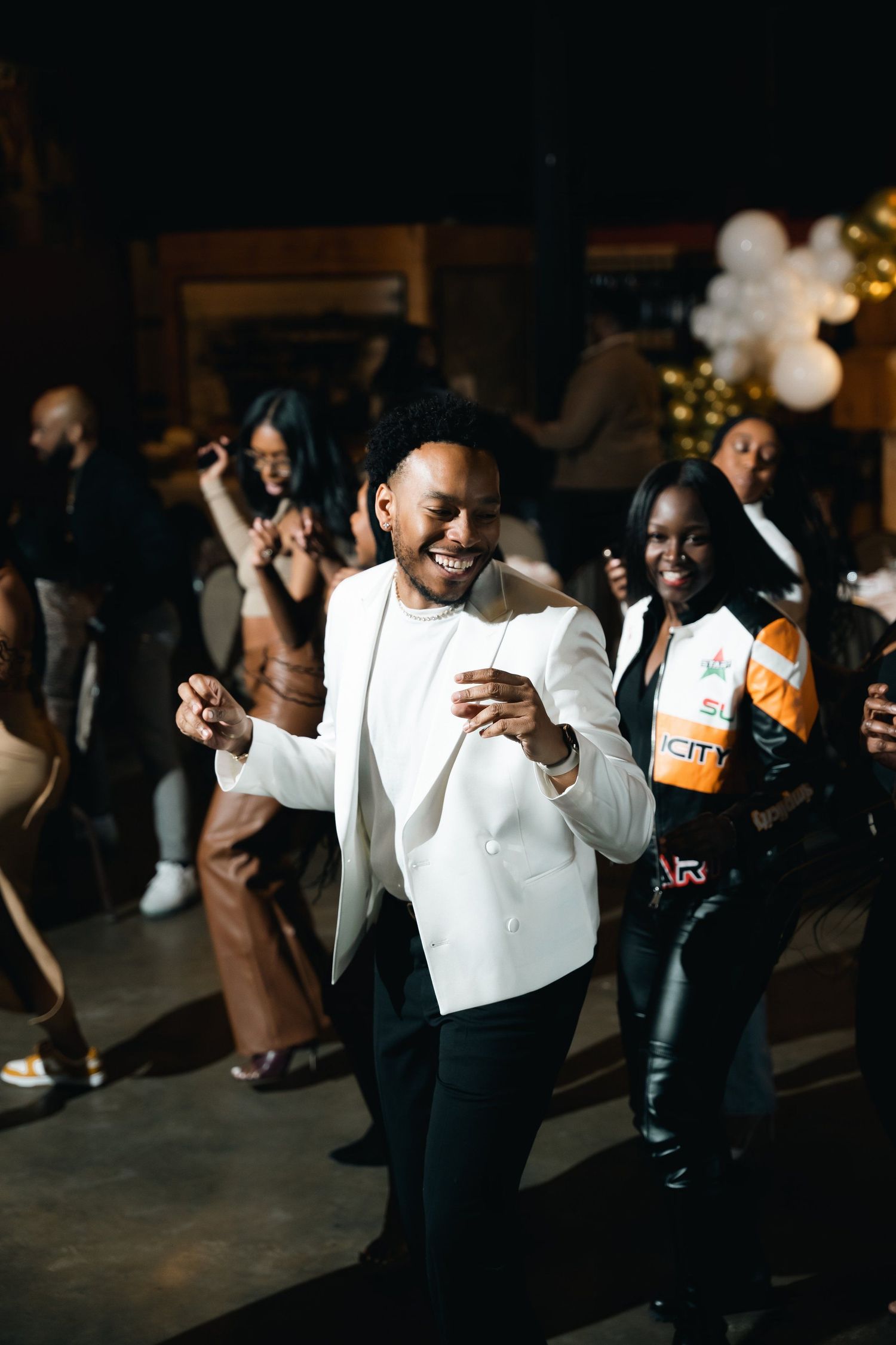 People dancing and celebrating at a lively party with someone in a white jacket in focus.