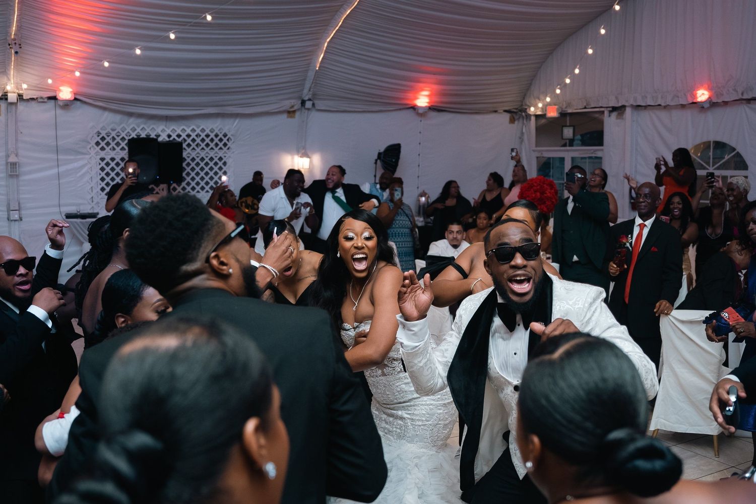 Wedding reception dance floor filled with guests celebrating under tent lighting.