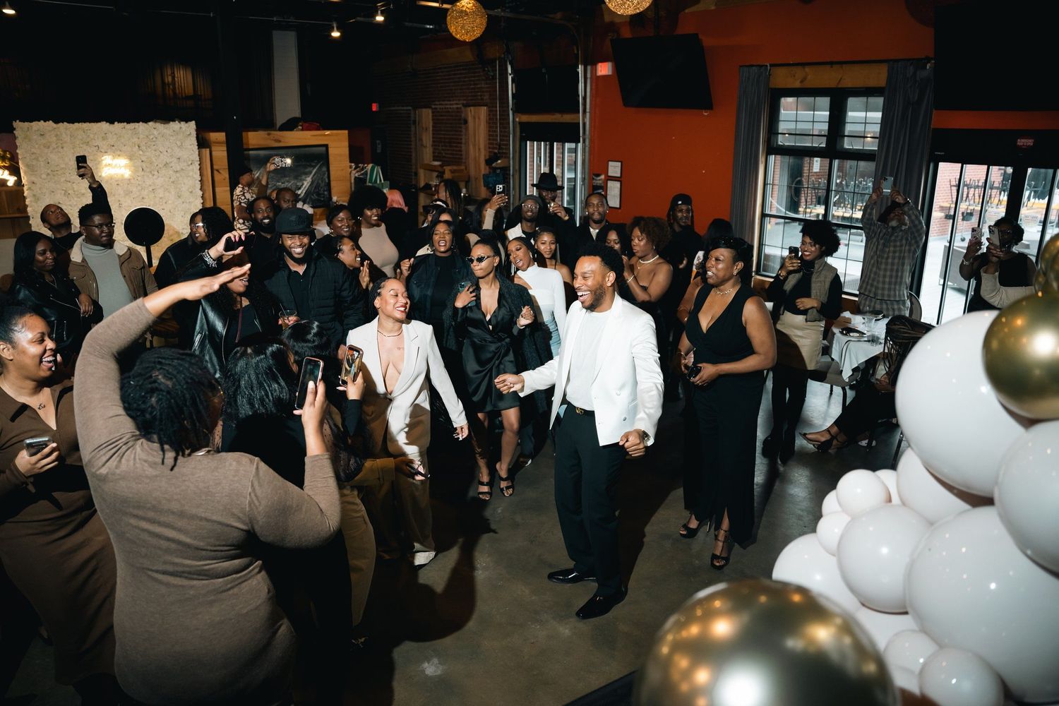 Large group dancing together at an indoor celebration with balloons and festive decor.