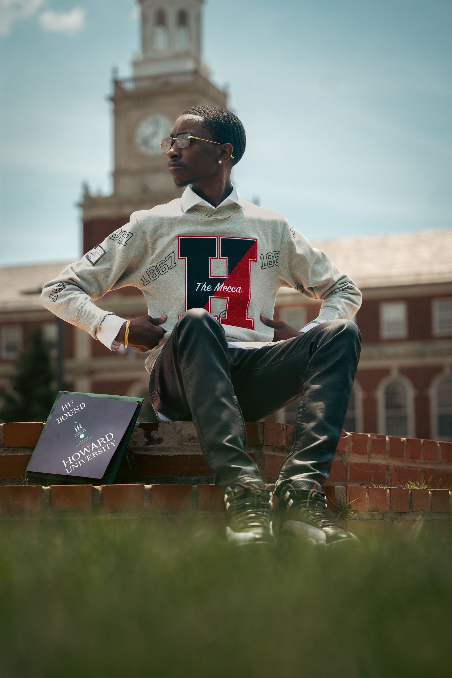 Student in varsity sweater poses on brick wall with historic clock tower in background.