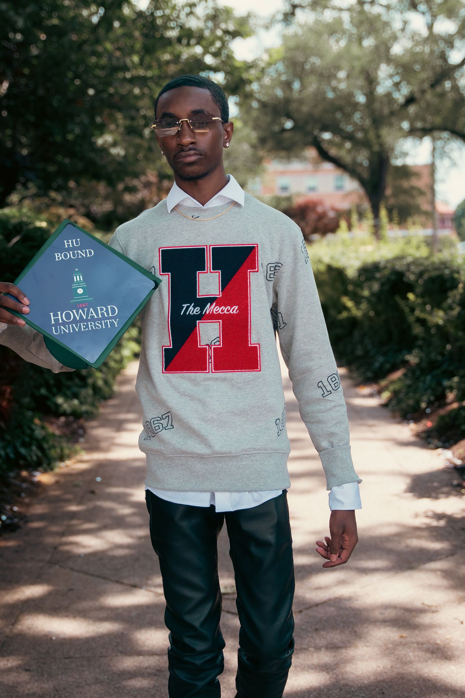 Student holding Howard University sign while wearing varsity sweater on campus pathway.
