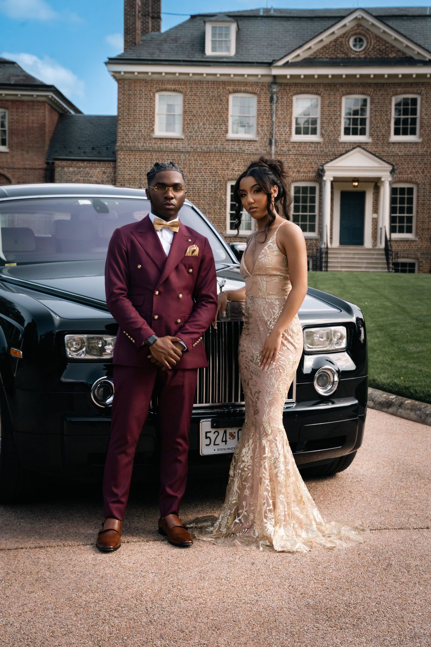 Elegant couple poses in formal attire next to a luxury Rolls Royce at a brick mansion.
