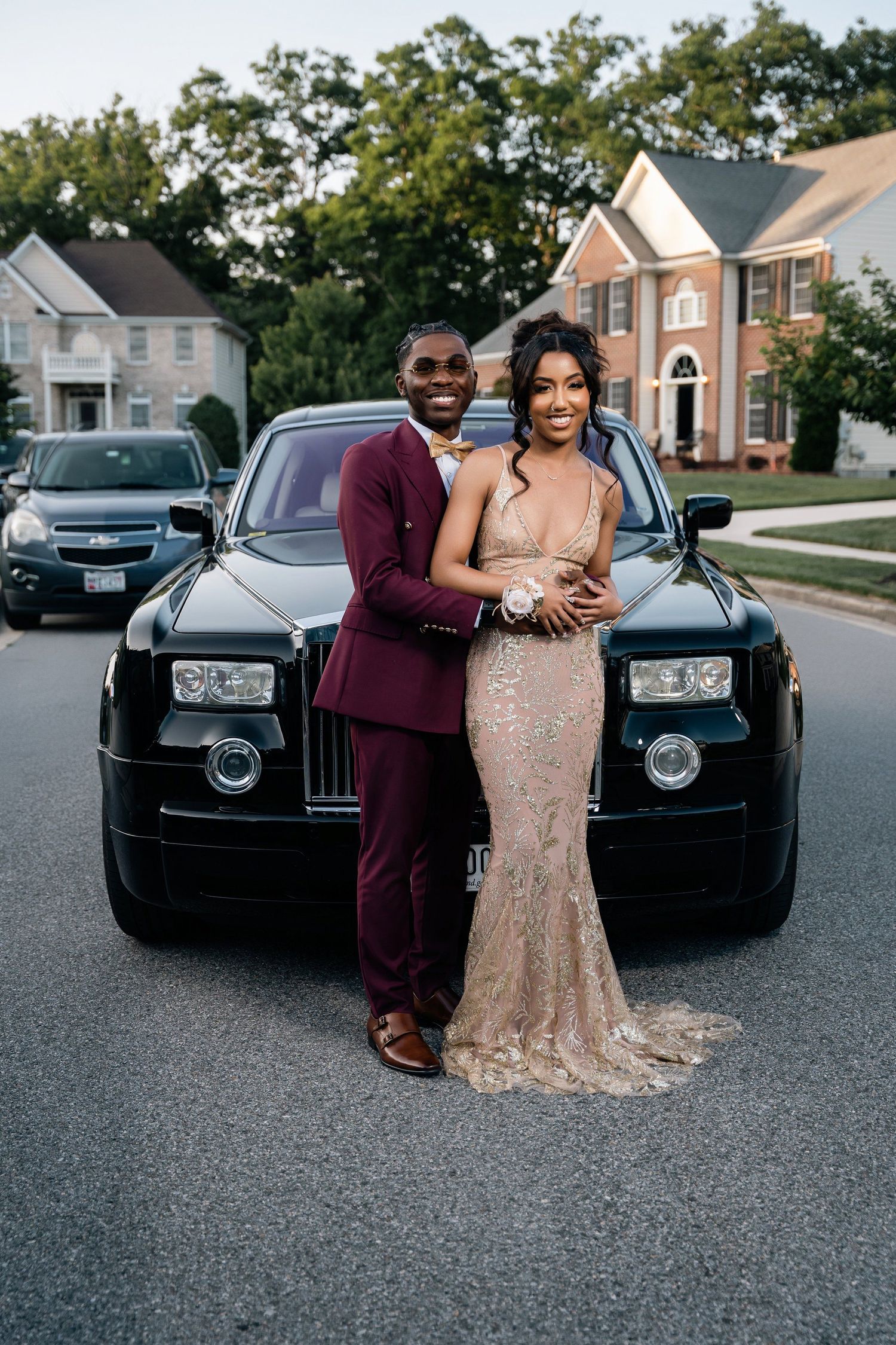 Formal couple in burgundy suit and champagne gown pose in front of a Rolls Royce and suburban homes.