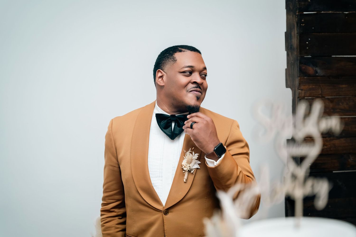 Person adjusting bowtie while wearing tan tuxedo jacket against white background.
