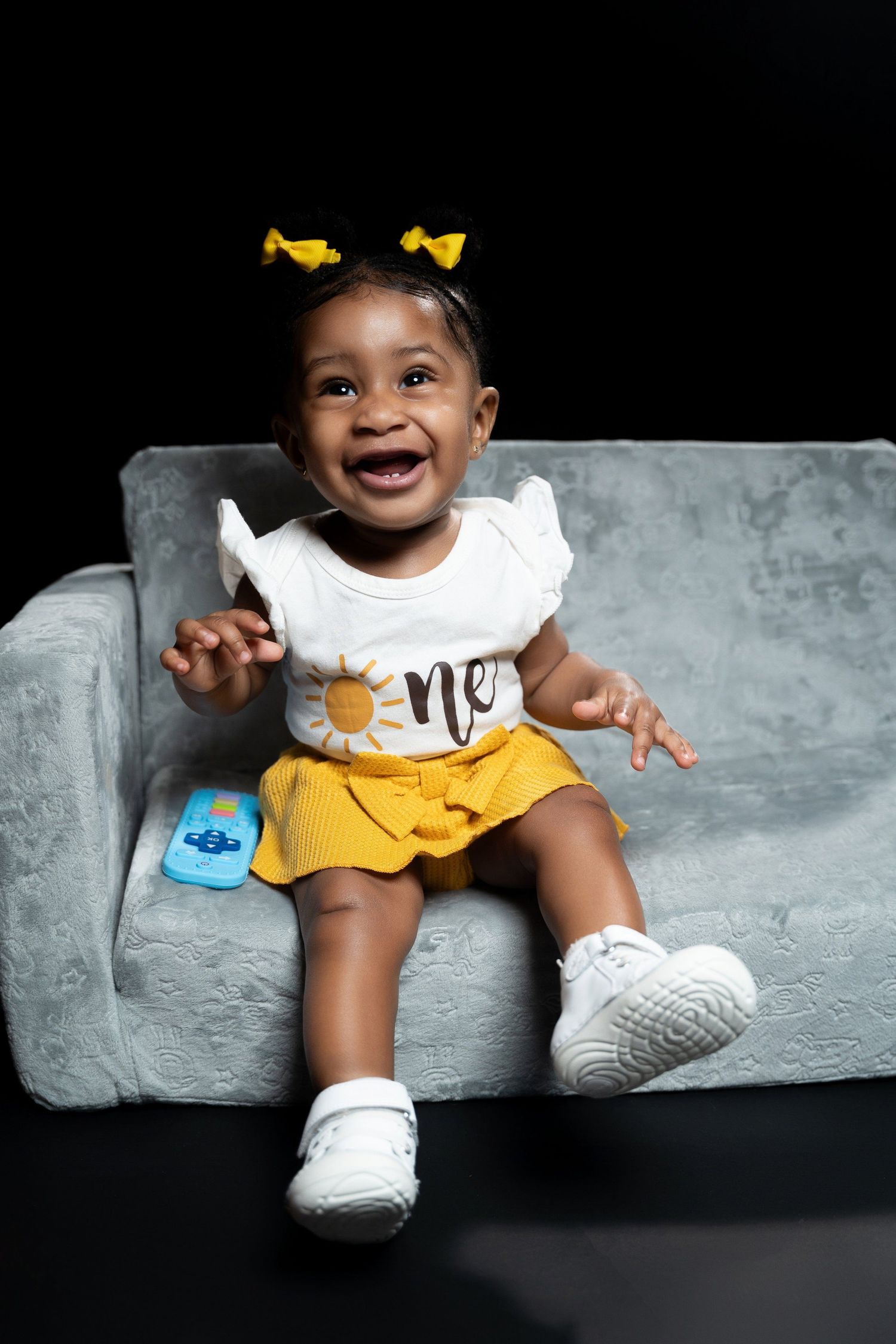 Smiling toddler in yellow and white outfit sits on gray chair with yellow hair accessories.