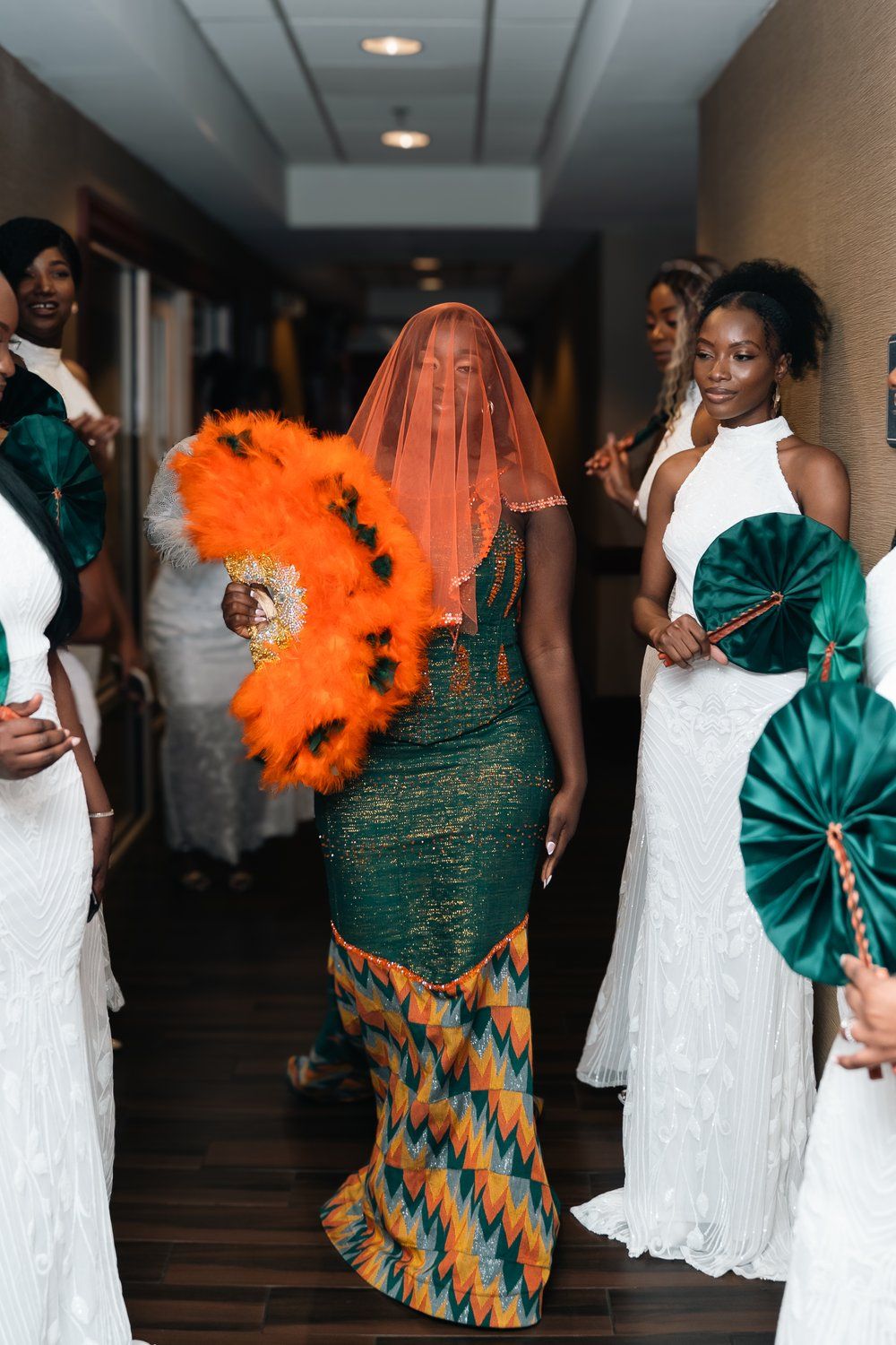 Wedding guests in formal attire with one wearing orange feathers and green traditional dress.
