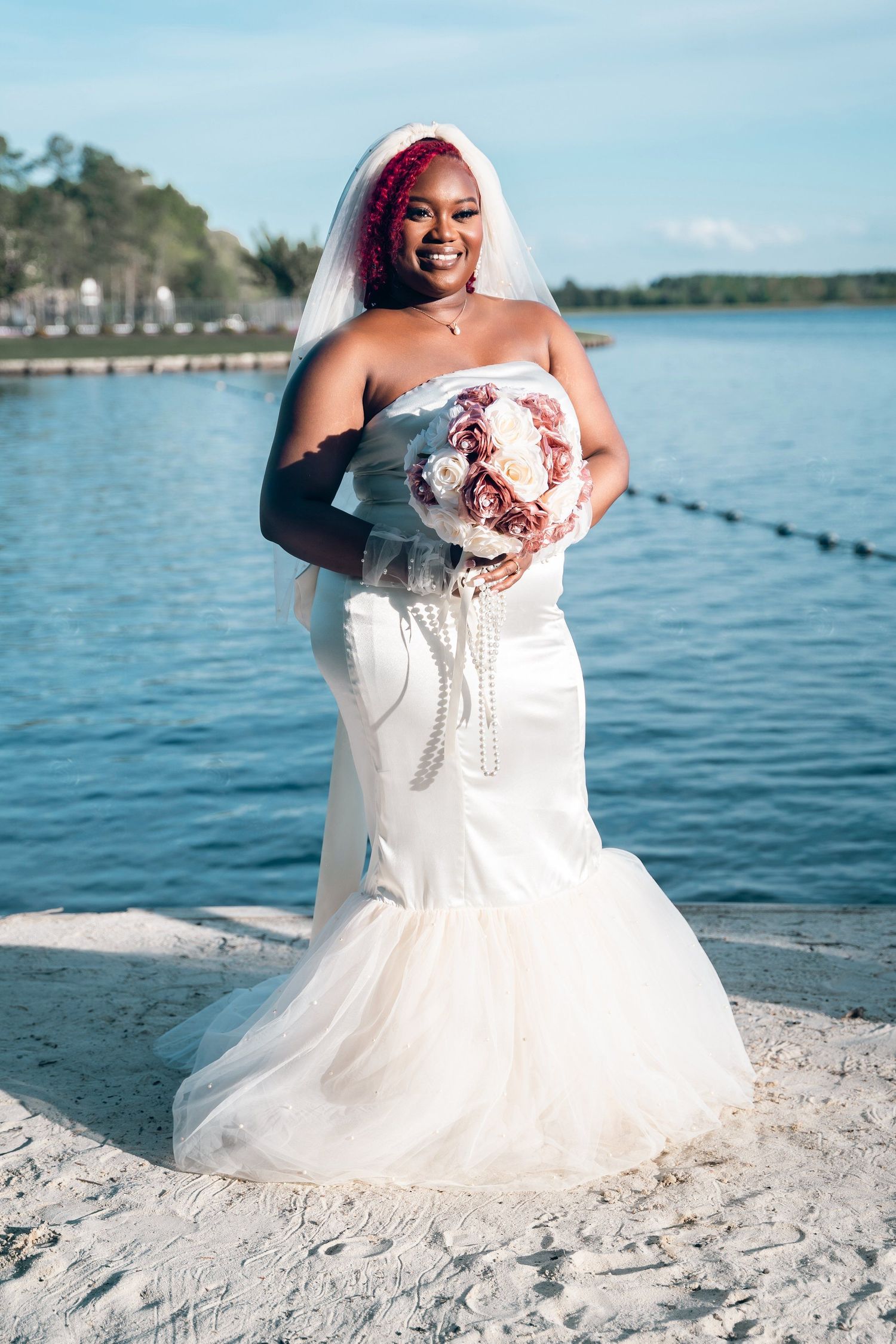 Bride in mermaid wedding dress holding bouquet poses on beach with water in background.