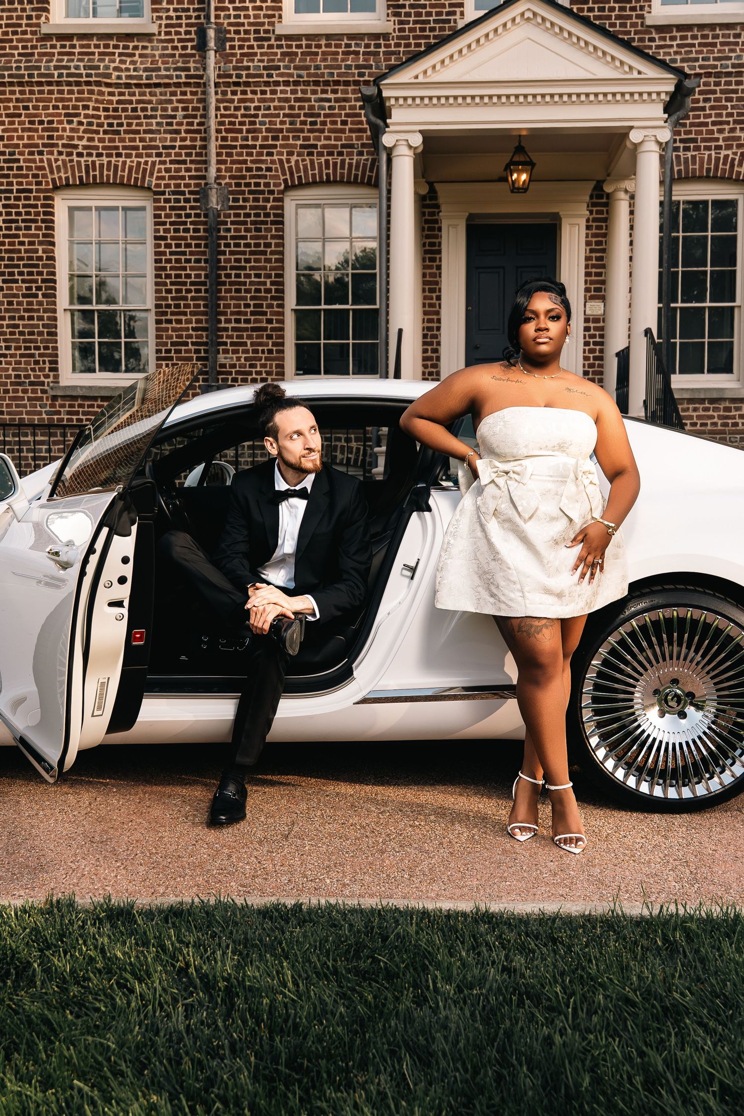 Wedding party poses playfully near classic white vintage car outside brick mansion venue.