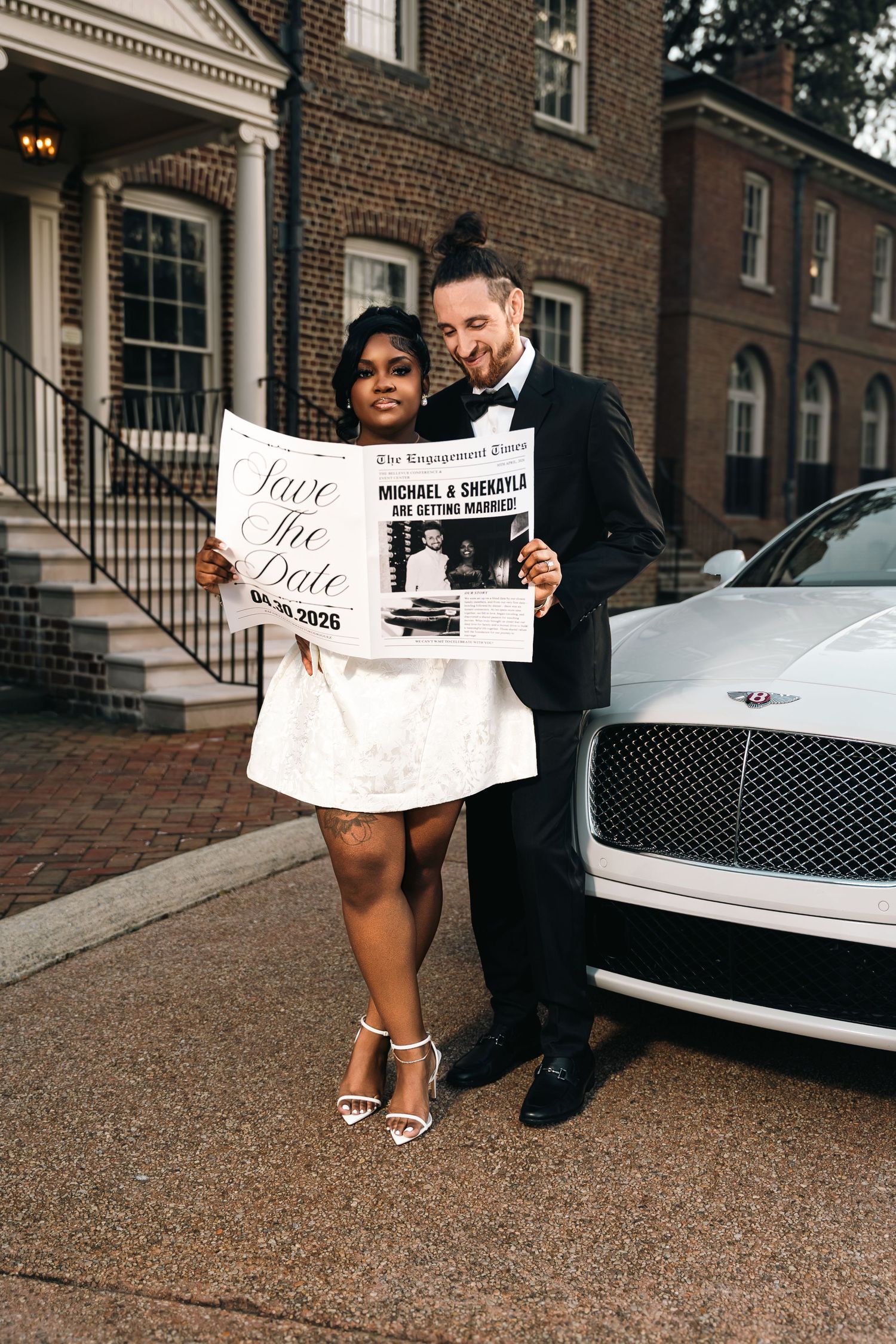 Two people pose with a newspaper in front of a brick building and luxury car on a cobblestone street.