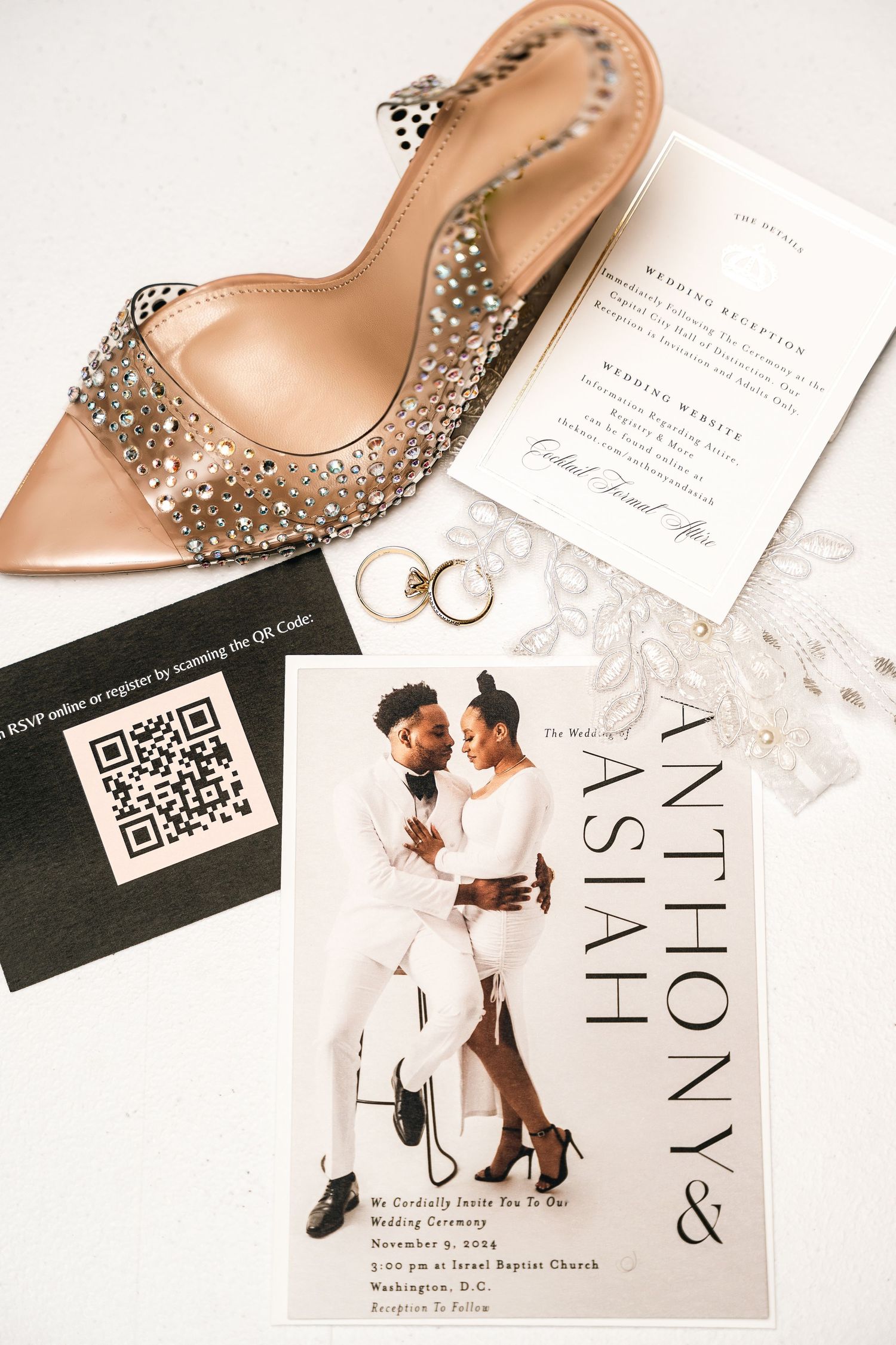 Wedding invitation with sparkly heels, rings and QR code laid out on white surface.