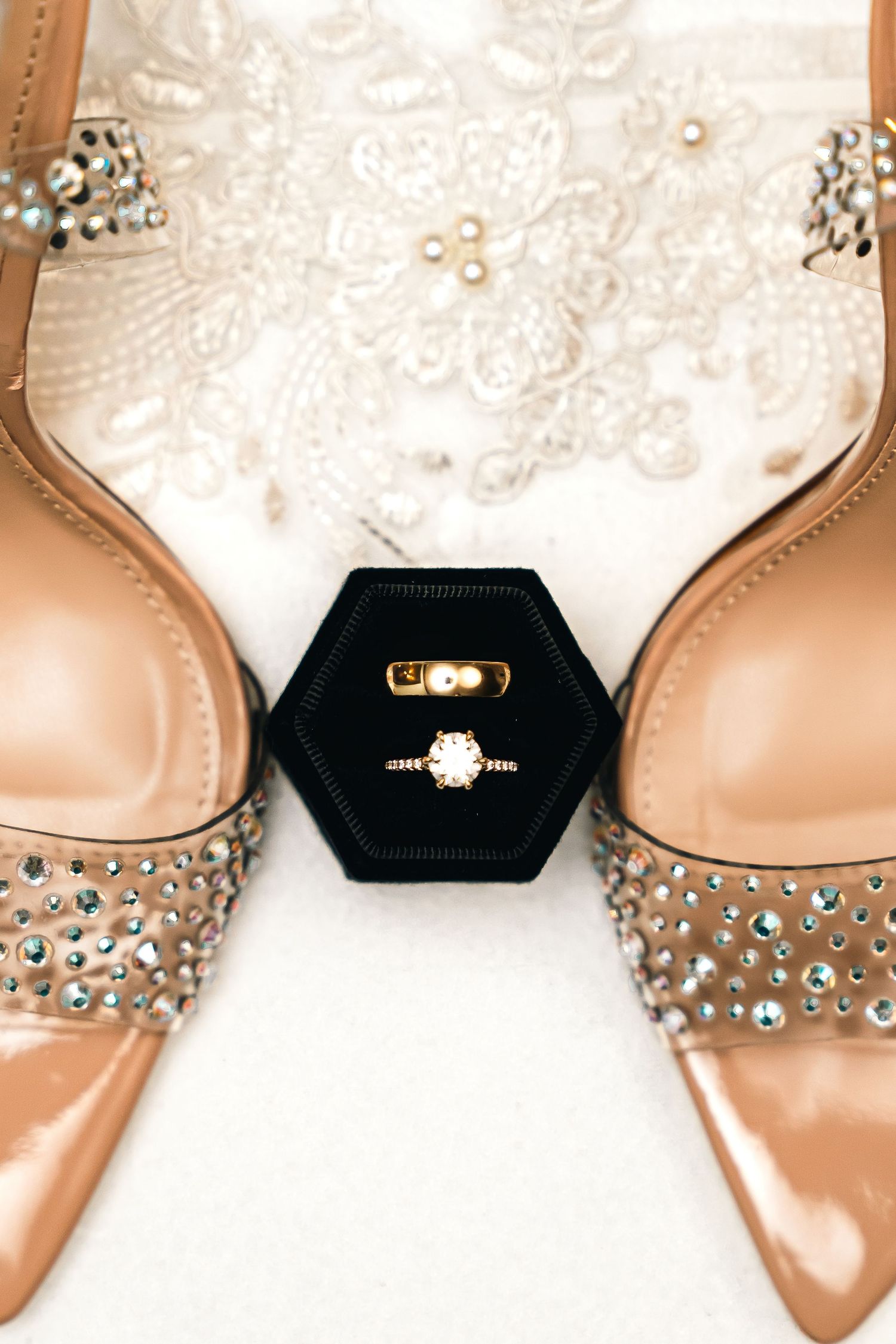Close-up of rhinestone-embellished nude heels with engagement rings on black display.