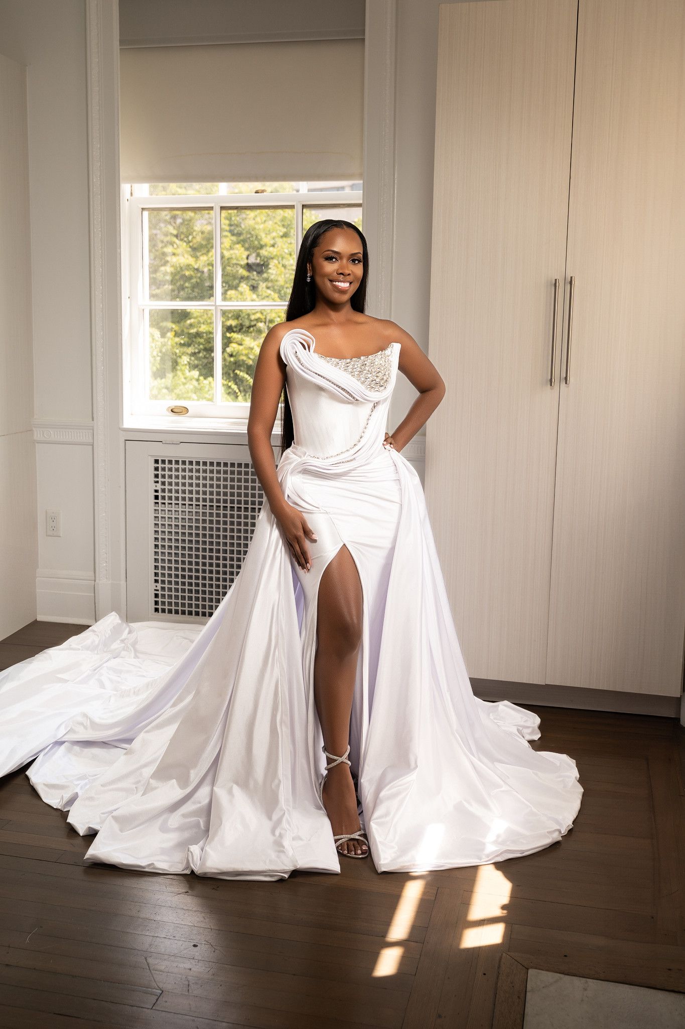 A flowing white wedding dress twirls elegantly across a hardwood floor in a sunlit room with white walls.