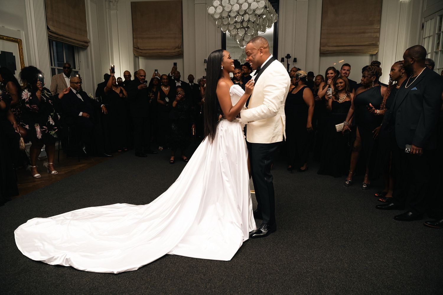 A bride in a flowing white wedding gown with a long train walks through an elegant venue filled with guests.