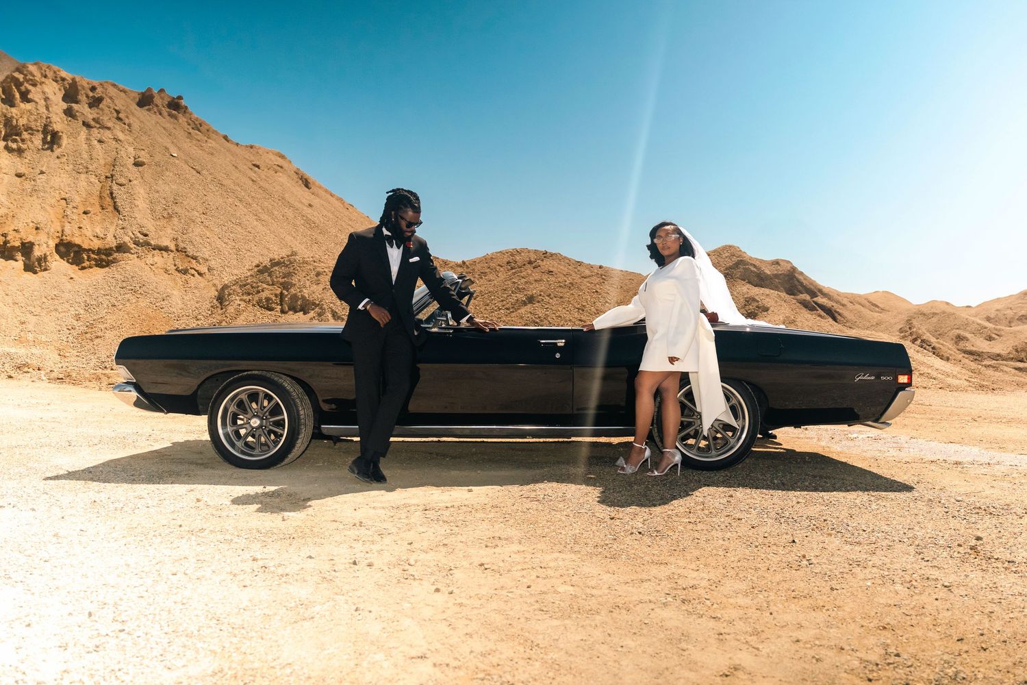 Two people in formal attire pose with a classic black convertible car in a desert landscape.