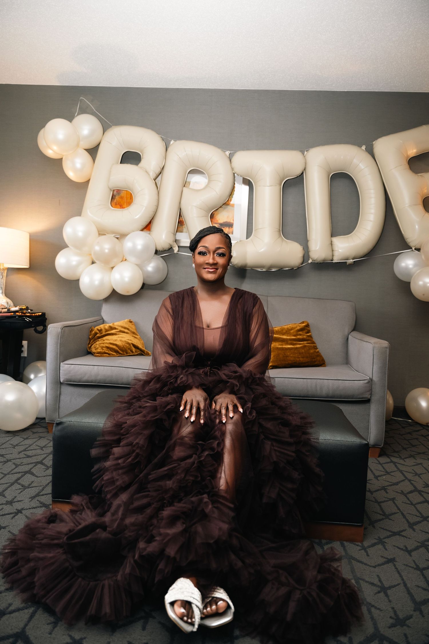 A person in a brown evening gown sits on a grey couch under white balloon letters spelling 'BRIDE' with elegant party decor.