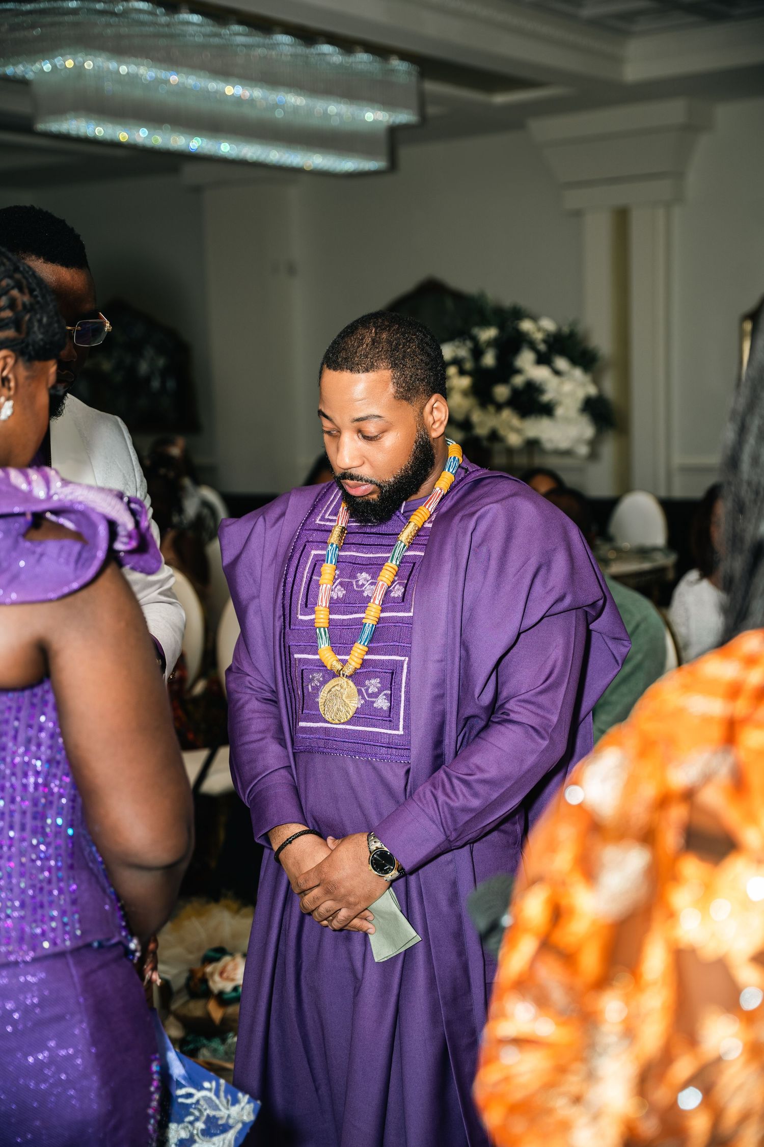 A person in a purple traditional African agbada with gold embellishments stands among other guests at a formal event.