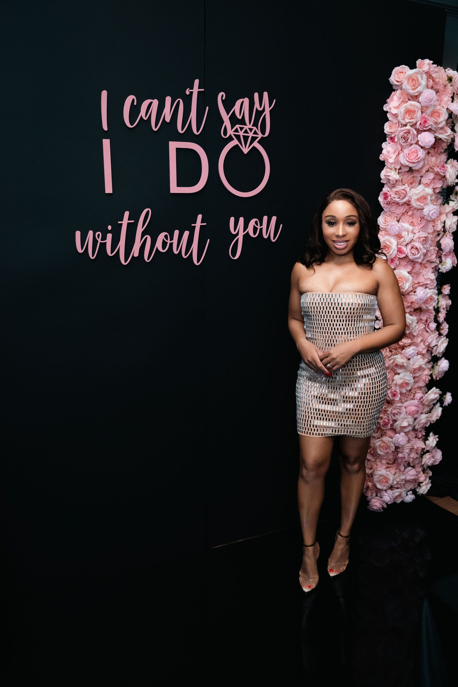 Woman in sparkly dress posing by wedding-themed wall decor with pink floral arrangements.