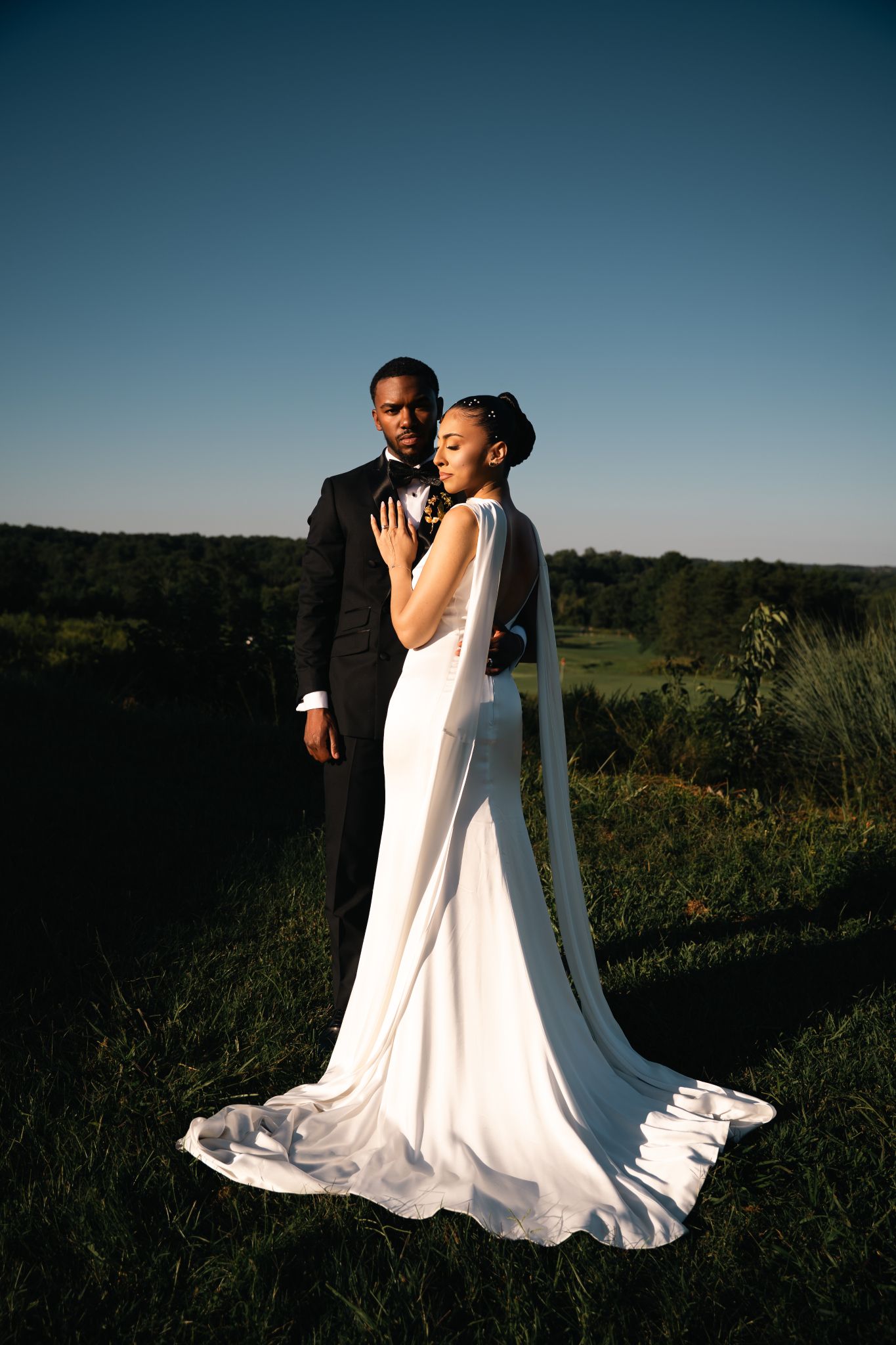Two brides in elegant white wedding dresses share a romantic moment in a sunlit meadow at sunset.
