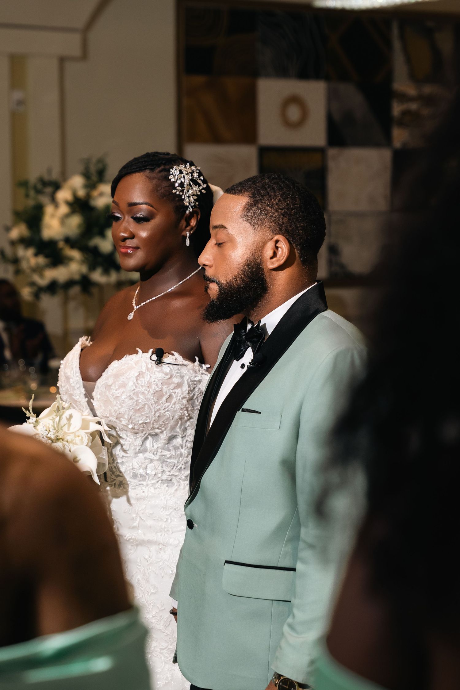 Newlyweds share intimate moment at wedding reception with bride in white lace dress and groom in mint green tuxedo.