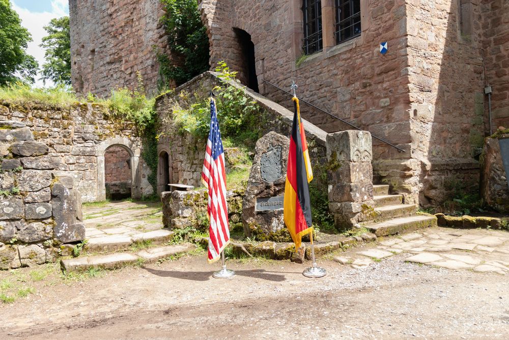 American and German flags displayed at the entrance of an ancient stone castle with stone steps and archways.