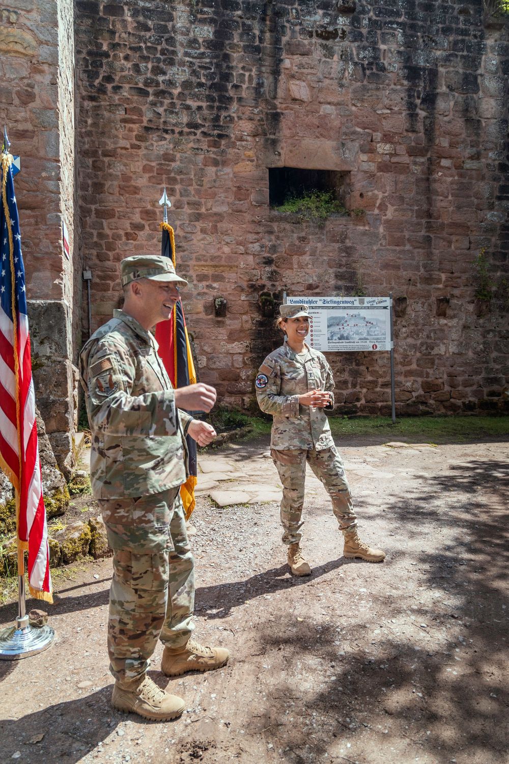 Military service members in camouflage uniforms stand near American flags next to an old stone wall on a sunny day.