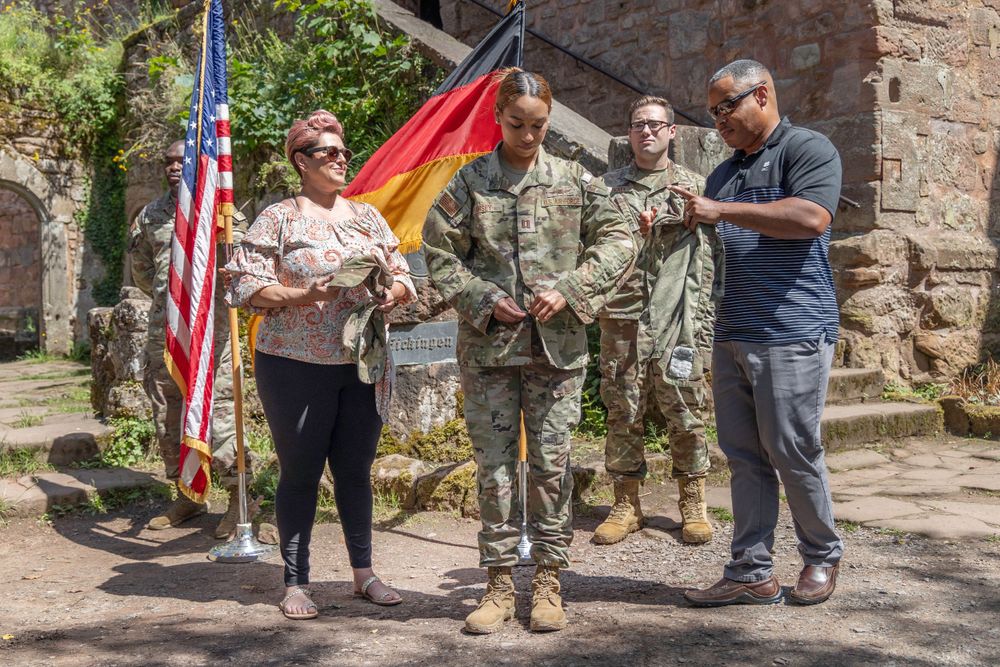 Military personnel stand with American and German flags outside a stone building while discussing.