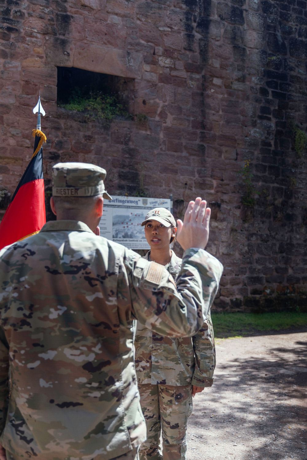 Military personnel in camouflage uniforms stand during an oath ceremony near historic stone walls.