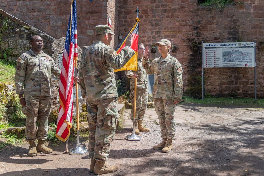 Military service members stand at attention with American and unit flags during a ceremony at a historic brick building.
