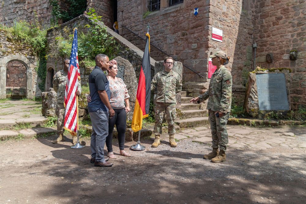 Military personnel and civilians stand together with American and German flags outside a historic stone building.