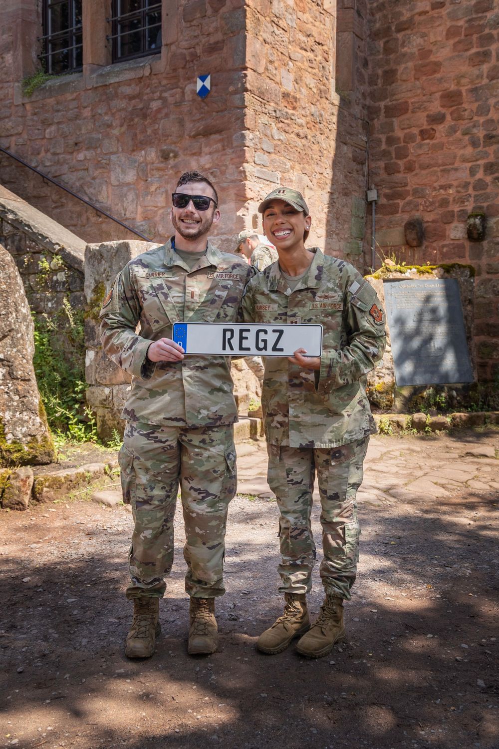 Two service members in camouflage uniforms stand holding a REGZ sign in front of a stone building on a sunny day.