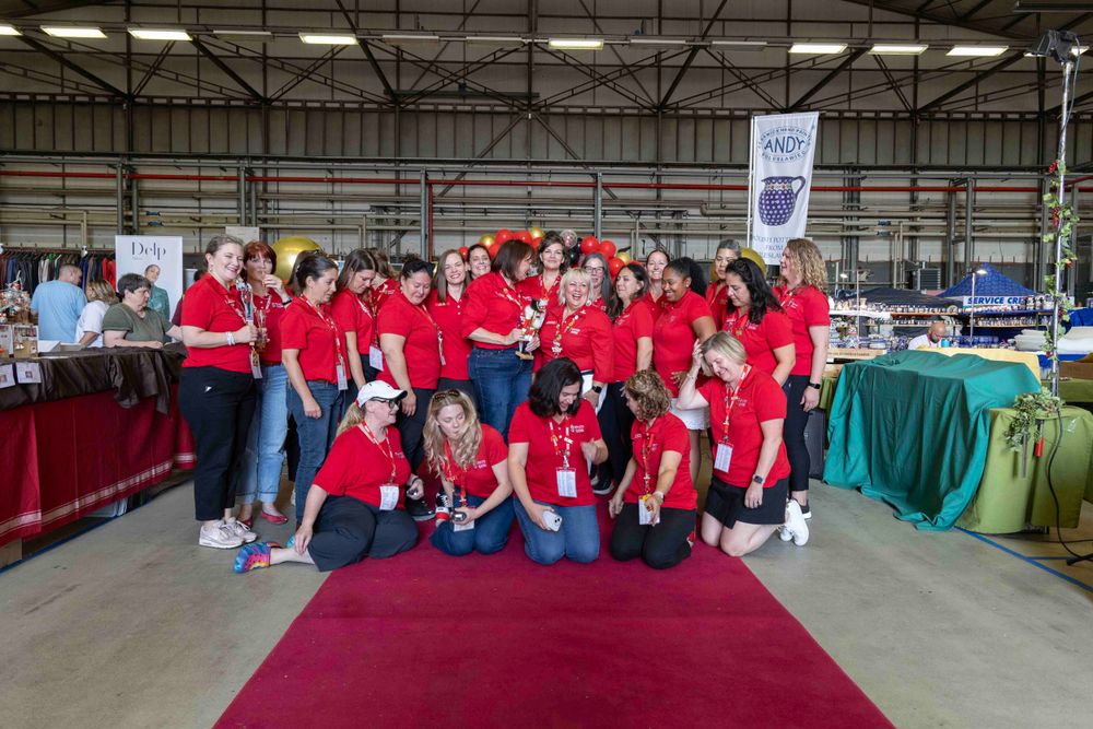 A group wearing matching red shirts poses together at an indoor event with vendor tables and displays.