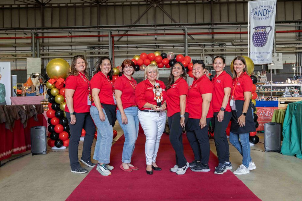 A group wearing matching red shirts poses together with gold and red balloons at an indoor event.