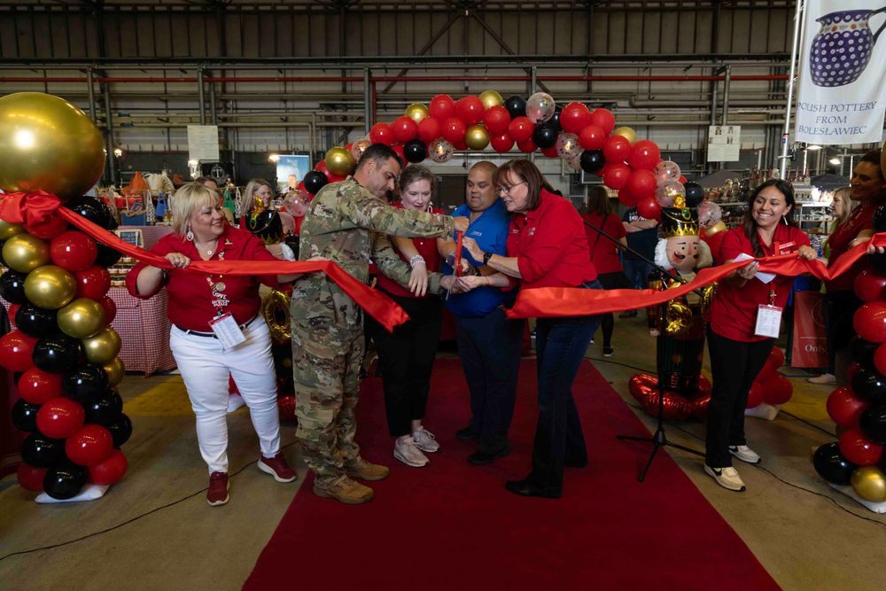 A group participates in a red ribbon cutting ceremony under red and gold balloon decorations at an indoor event.