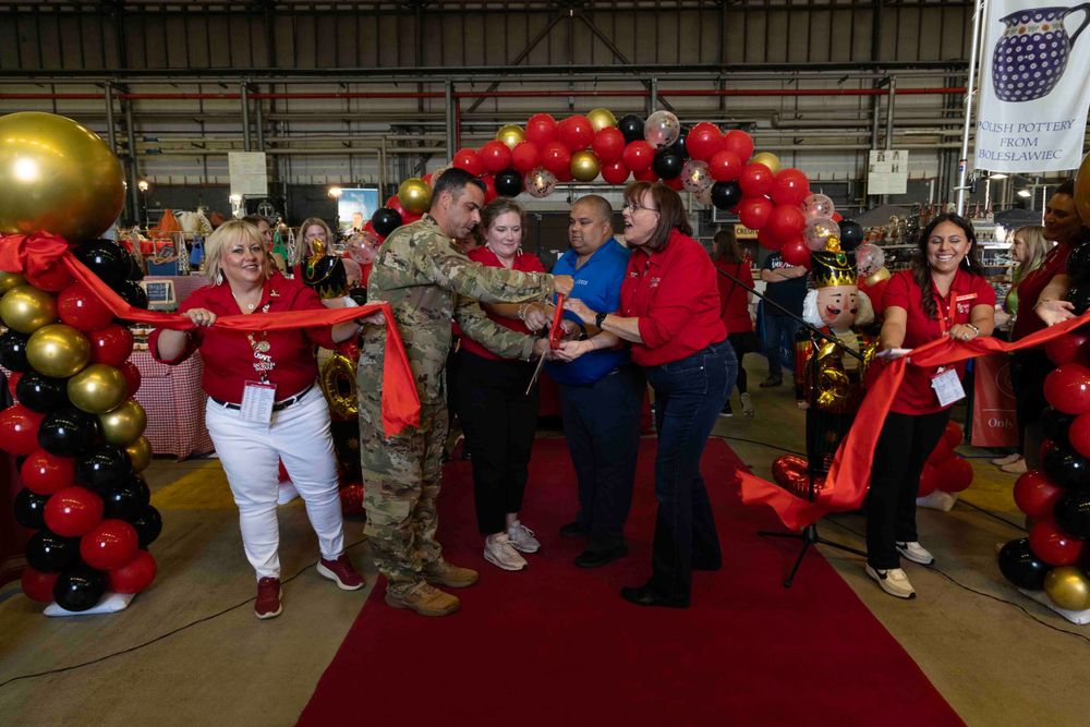 A group celebrates at an event under a festive red and black balloon arch on a red carpet in what appears to be a hangar.