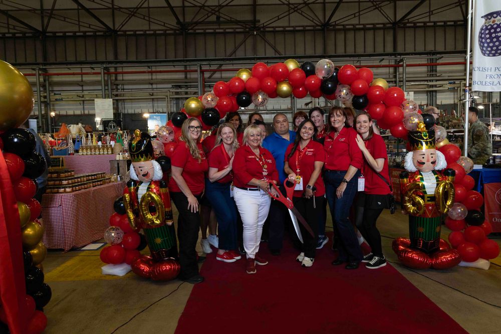 Group photo under a festive red balloon arch at an indoor event with people wearing red and blue shirts on a red carpet.