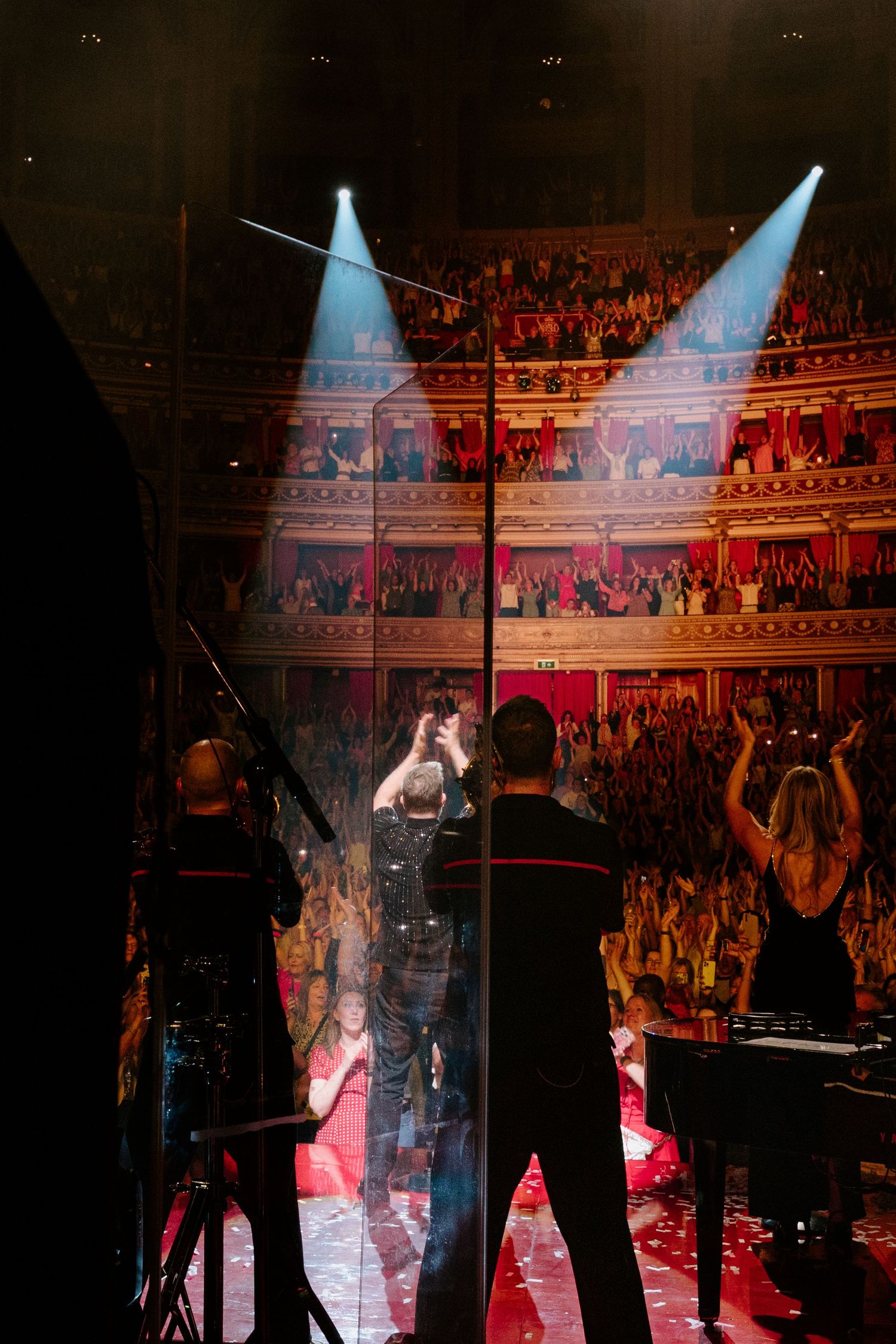 Concert performers silhouetted on stage under dramatic blue and red spotlights in front of energetic crowd.
