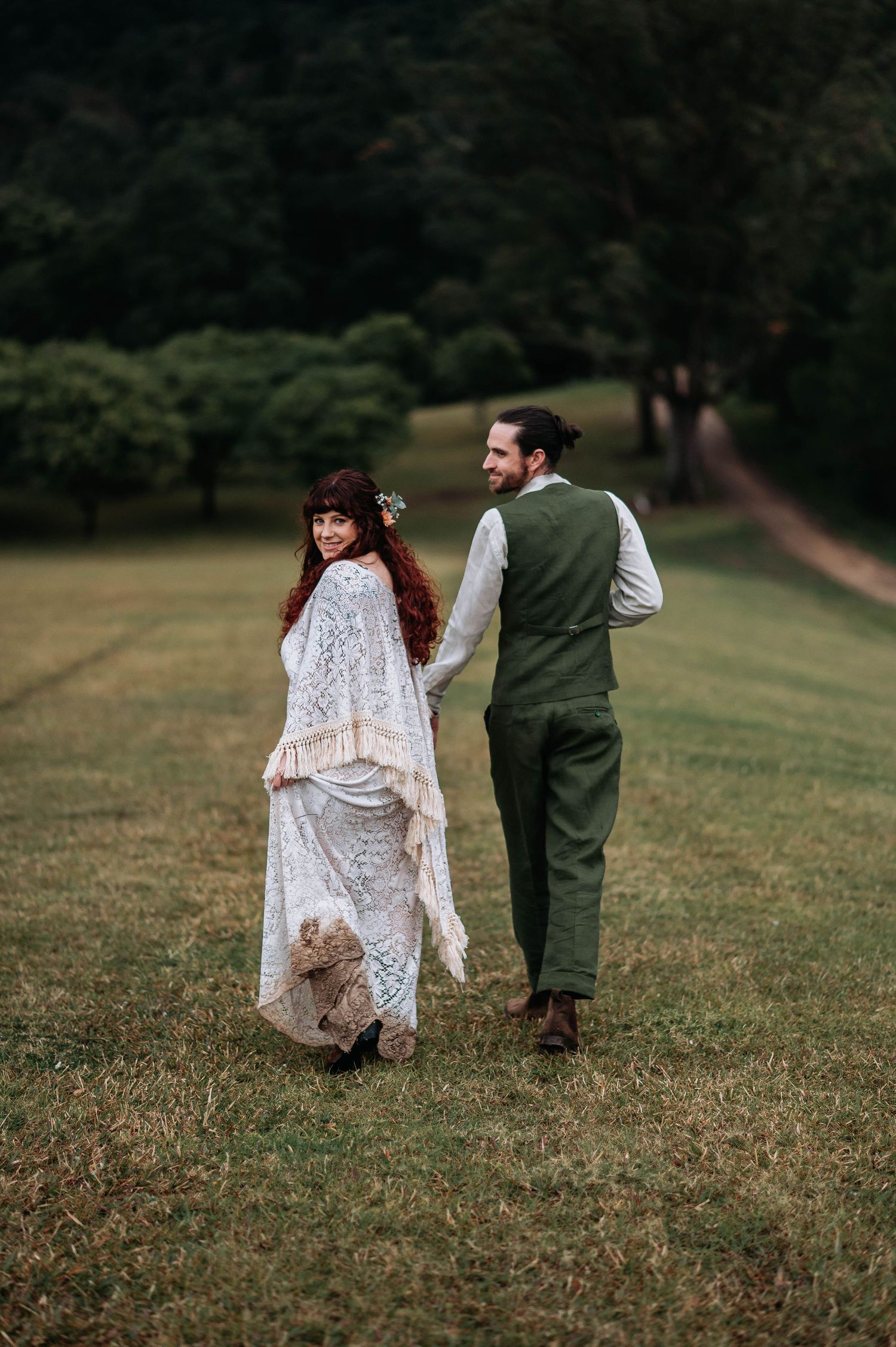 A couple in vintage-inspired wedding attire walks hand in hand across a grassy meadow on an overcast day.