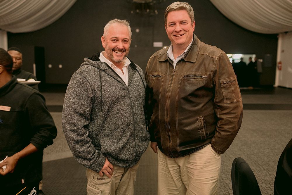 Two smiling men posing together at an indoor corporate event, one wearing a grey sweater and the other a brown leather jacket.