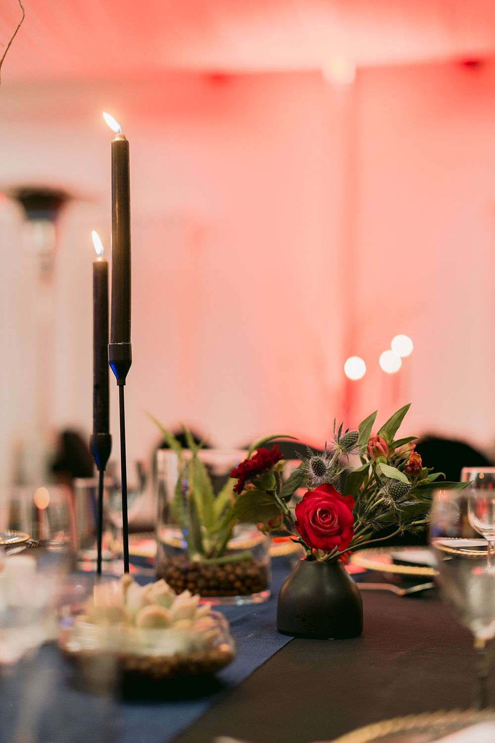 Elegant dinner table centerpiece with tall black candles and a red rose arrangement in a dark vase at a gala event.