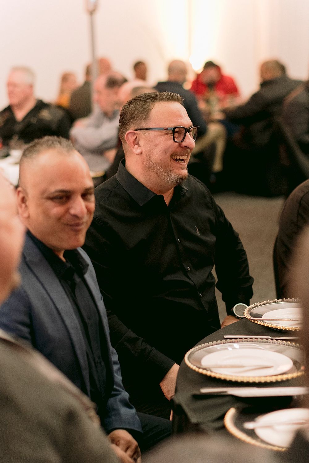 Two smiling men in black attire seated at an elegantly set dinner table at a formal event.
