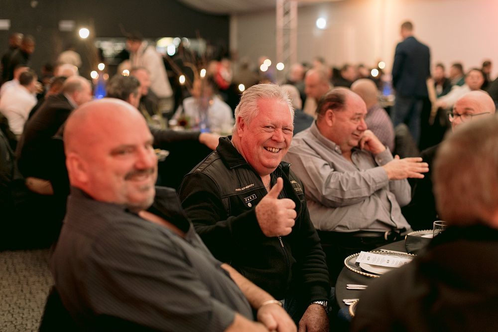 Group of men laughing at a corporate banquet, one giving a thumbs up in a crowded event venue.