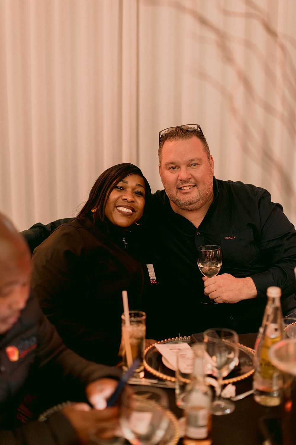 Smiling couple posing together at a formal dinner event, man holding a glass of white wine.