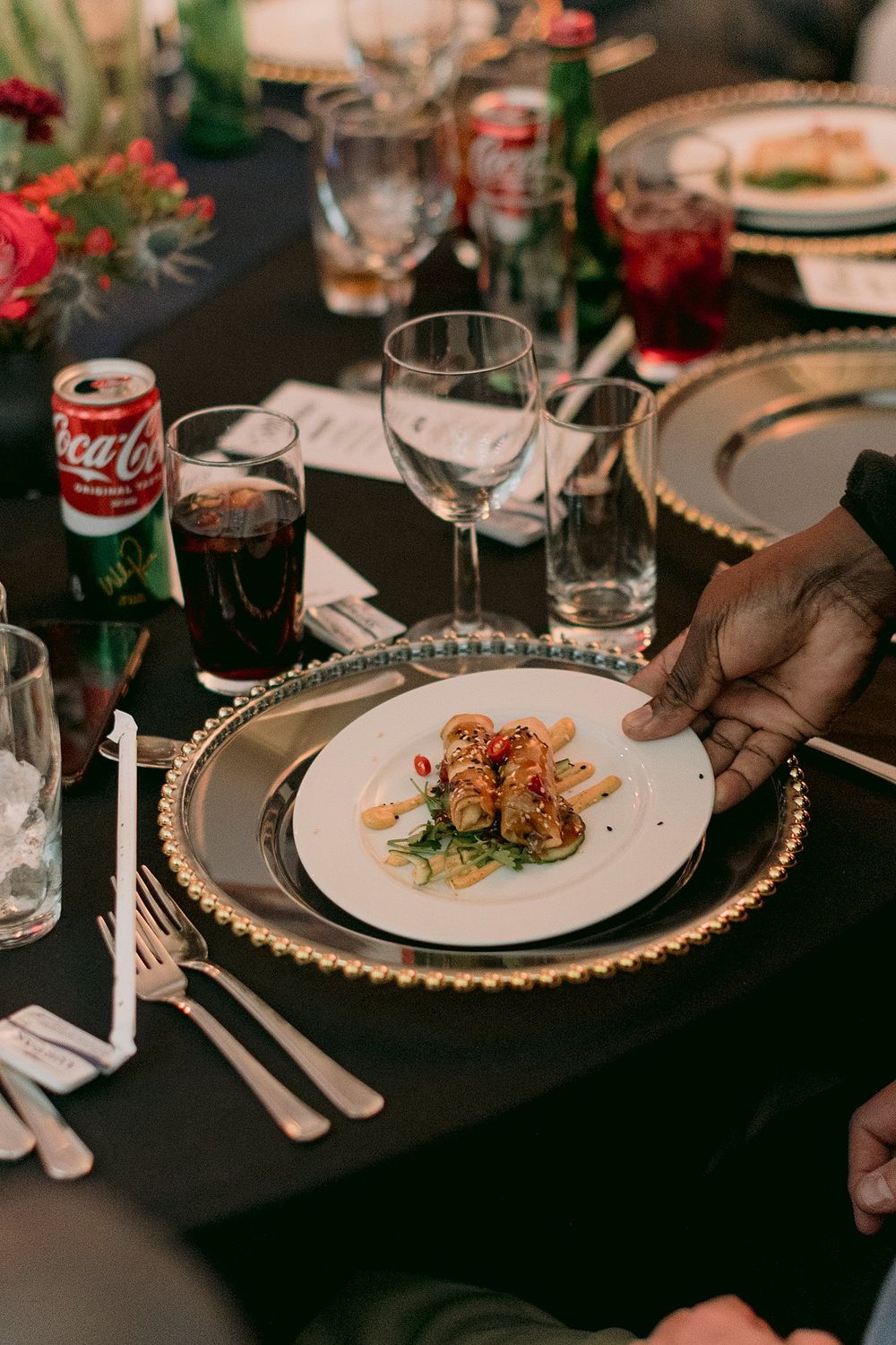 Hand placing an elegantly plated starter dish on a gold-beaded charger at a formal dinner table.