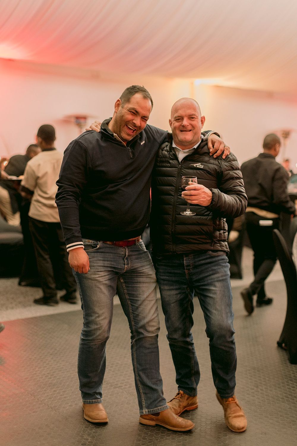 Two smiling men posing together at an indoor corporate evening event, one holding a drink.