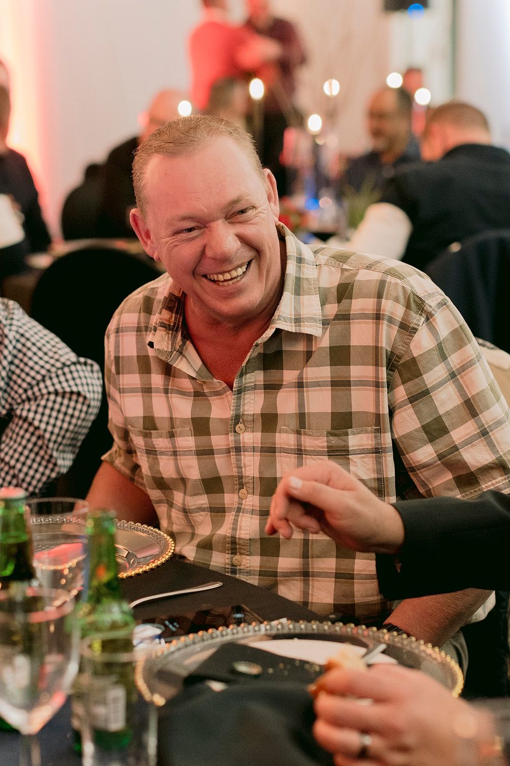 Smiling man in plaid shirt laughing at a formal dinner event seated at a table.