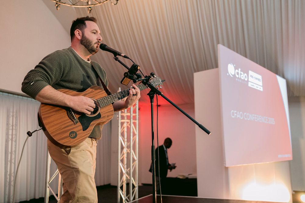 Male guitarist performing on stage at a CFAO conference event with a projection screen behind him.