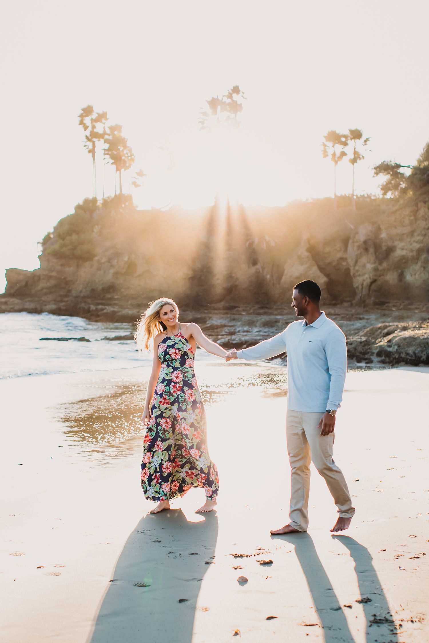 Couple playfully dancing together on a sunny beach at sunset near rocky coastal cliffs.